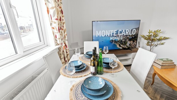 A dining area with a table set for four, featuring blue plates and wine glasses. A television displaying 'Monte Carlo Monaco' is mounted on the wall.