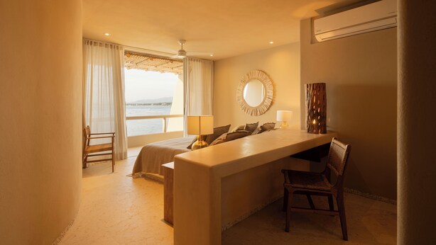 Habitación de hotel con cama, escritorio, silla, espejo redondo y vista al mar.