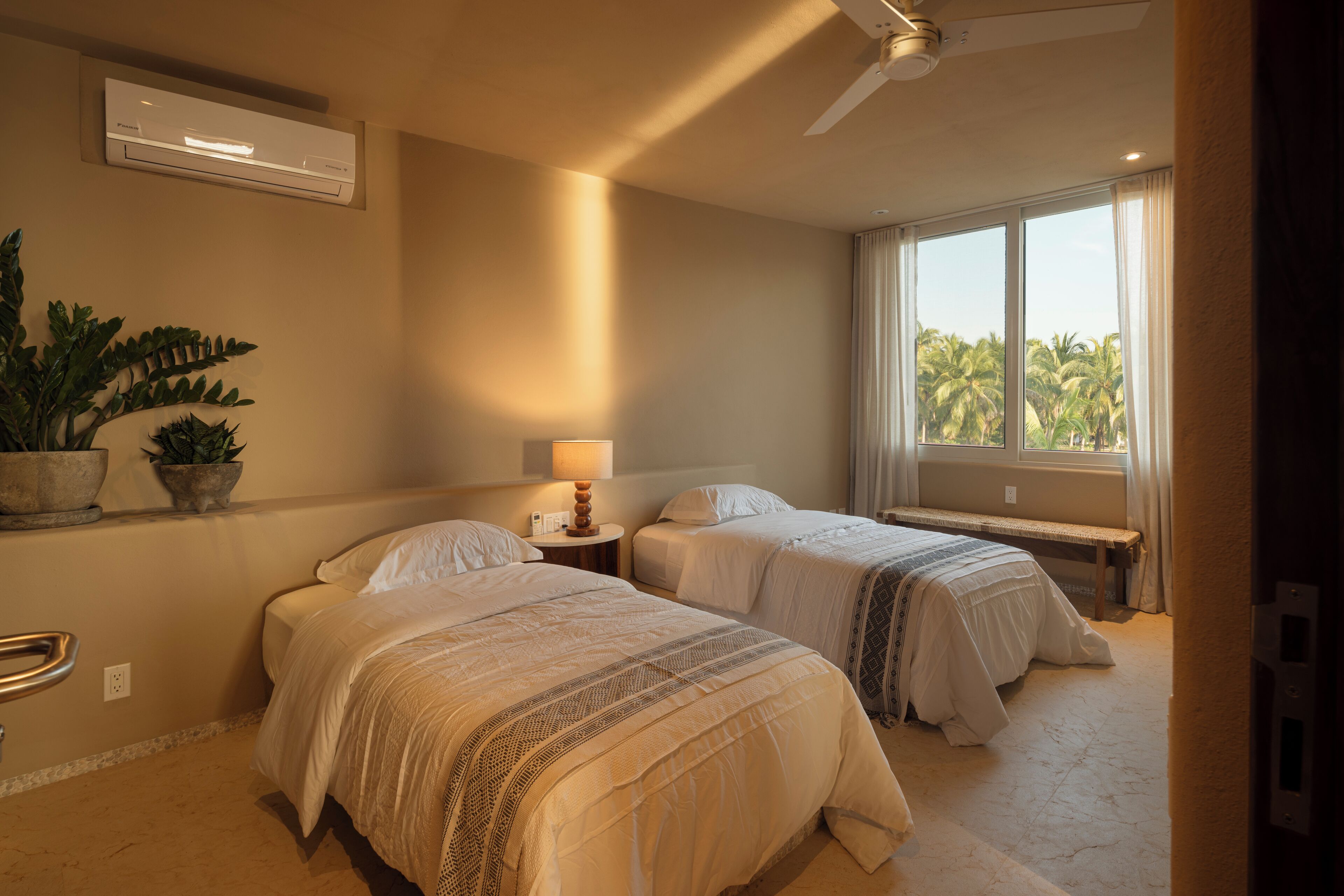 Ein Hotelzimmer mit zwei Betten, einem Deckenventilator, einer Klimaanlage, einer Nachttischlampe und einem Fenster mit Blick auf Palmen.