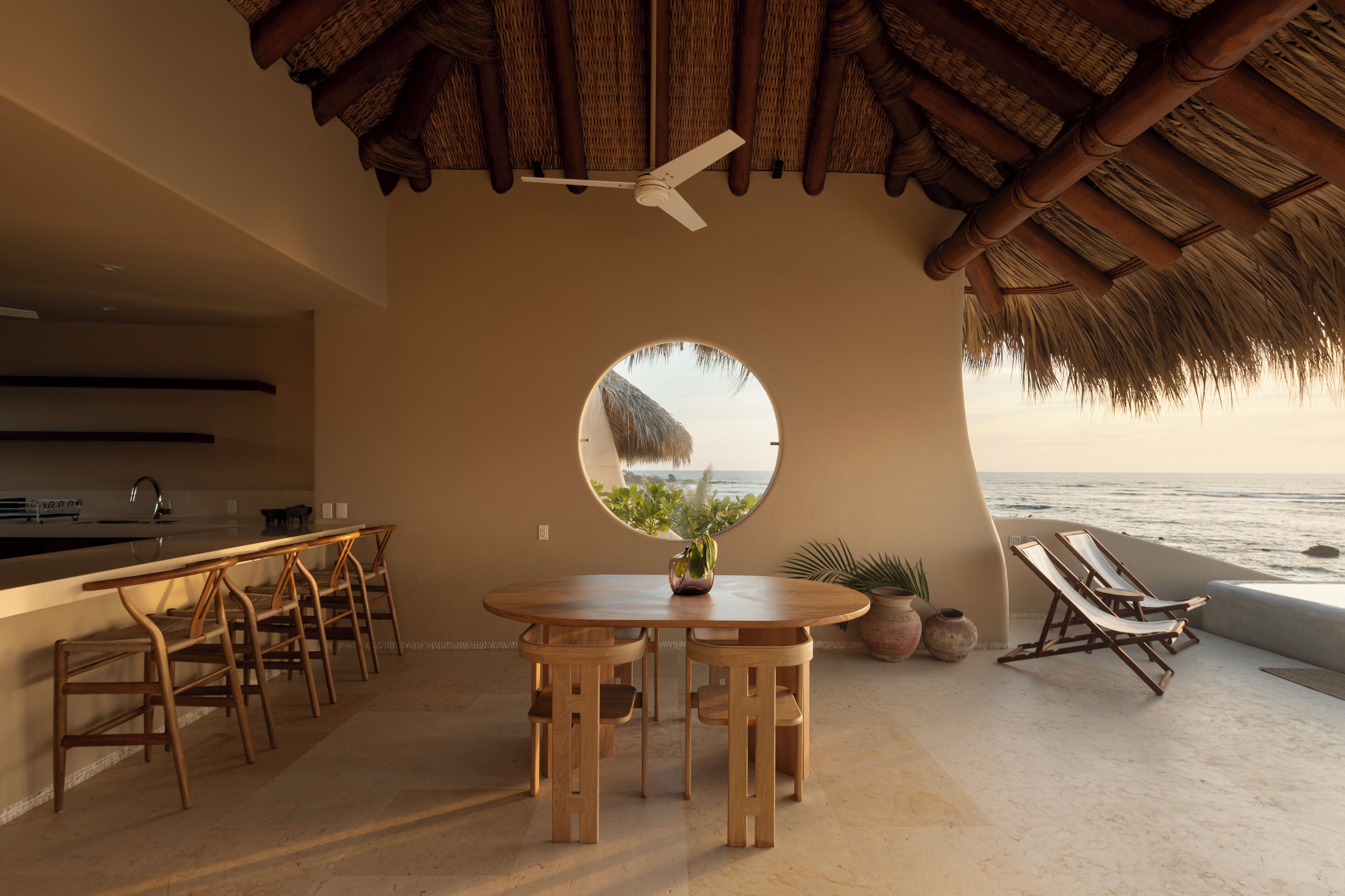 A beachside dining area with a round table, wooden chairs, a ceiling fan, and a view of the ocean through a circular window.