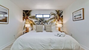 A bedroom with a large bed, two bedside lamps, a window, and a tropical mural on the wall.