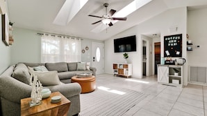 A modern living room with a grey sofa, a wooden coffee table, and a ceiling fan. There is a TV mounted on the wall and a coffee station with a sign that reads "Coffee and Tea".