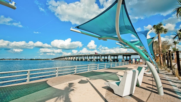A waterfront promenade with a bridge in the background, a metal sculpture, and palm trees.