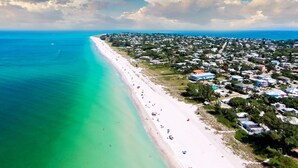 An aerial view of a coastal area with a long sandy beach, a dense residential area with houses, and a clear blue sea.