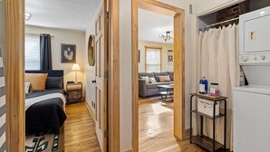 A compact room with a bed, bedside table, lamp, and a small kitchenette area with a refrigerator and a laundry station.