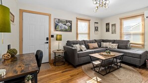 A living room with a grey sofa, a wooden coffee table, and a rug. There is a door, a lamp, and a wall-mounted picture.