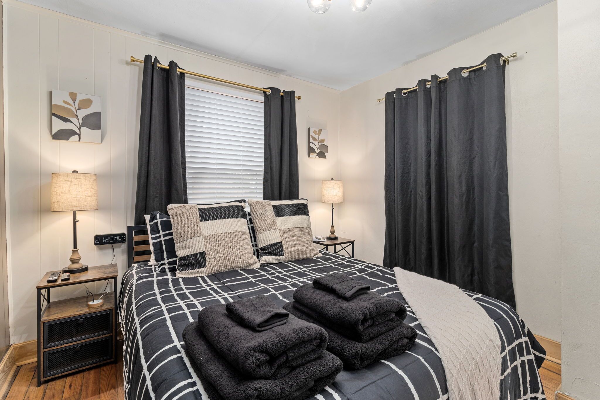 Ein ordentlich bezogenes Bett mit einem schwarz-weißen Karokissen, zwei Kissen und zwei gefaltete Handtücher. Zwei Nachttische mit Lampen, einer Digitaluhr und einem kleinen Lautsprecher stehen bereit. Das Zimmer verfügt über ein Fenster mit Jalousien und zwei an der Wand befestigte Kunstwerke.