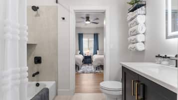 Ein modernes Badezimmer mit Badewanne, Toilette und Dusche. Ein Gang führt zu einem Schlafzimmer mit Bett und Nachttisch.