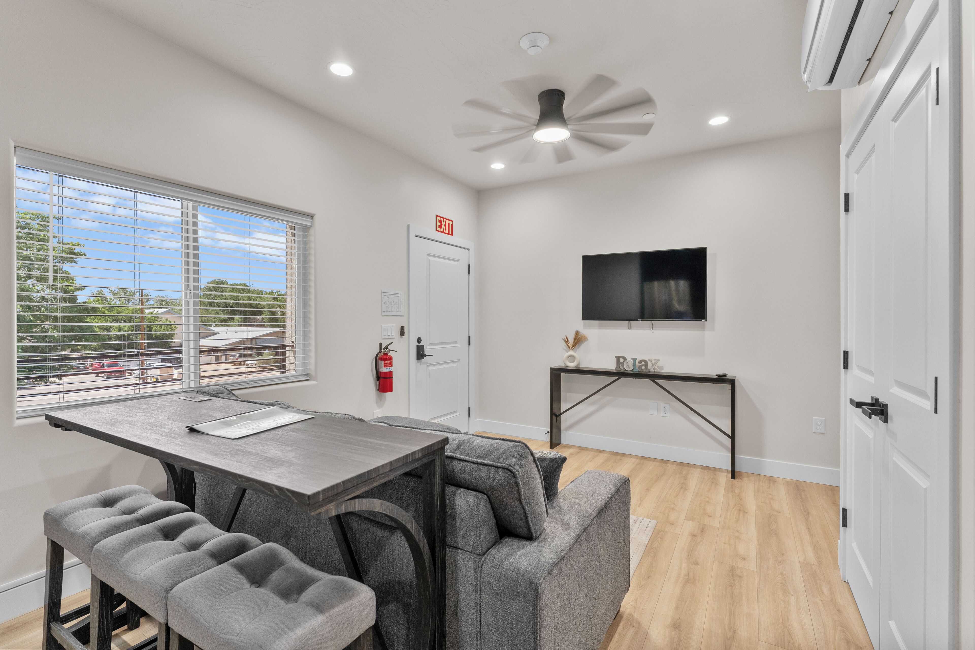 A modern kitchen with a dining area, a TV mounted on the wall, and a window with blinds.