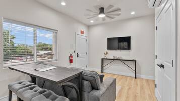 A modern kitchen with a dining area, a TV mounted on the wall, and a window with blinds.