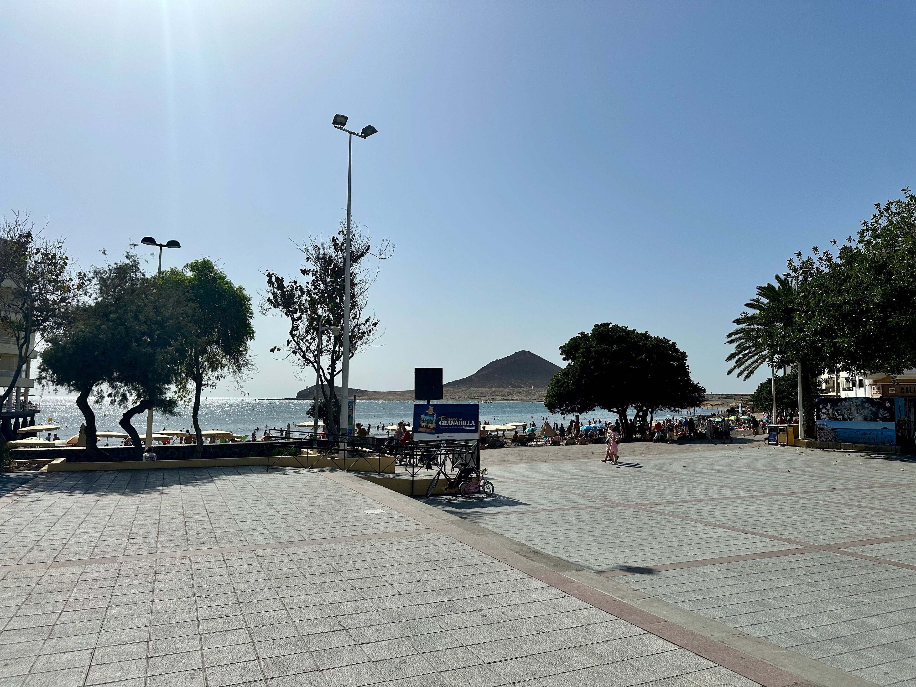 Paseo pavimentado con árboles y un cartel, con vistas a una playa donde hay gente y una montaña al fondo.