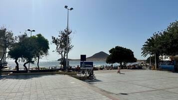 Paseo pavimentado con árboles y un cartel, con vistas a una playa donde hay gente y una montaña al fondo.