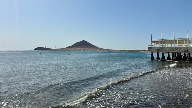 Escena costera con cielo despejado, una montaña al fondo y un muelle que se adentra en el agua.