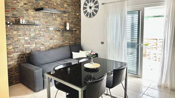 A modern kitchen with a brick wall backsplash, a black dining table with chairs, a gray sofa, and a large wall clock. - Pambnb - House next to the Beach, Terrace, Fiber Wi-Fi, BBQ (Abades)