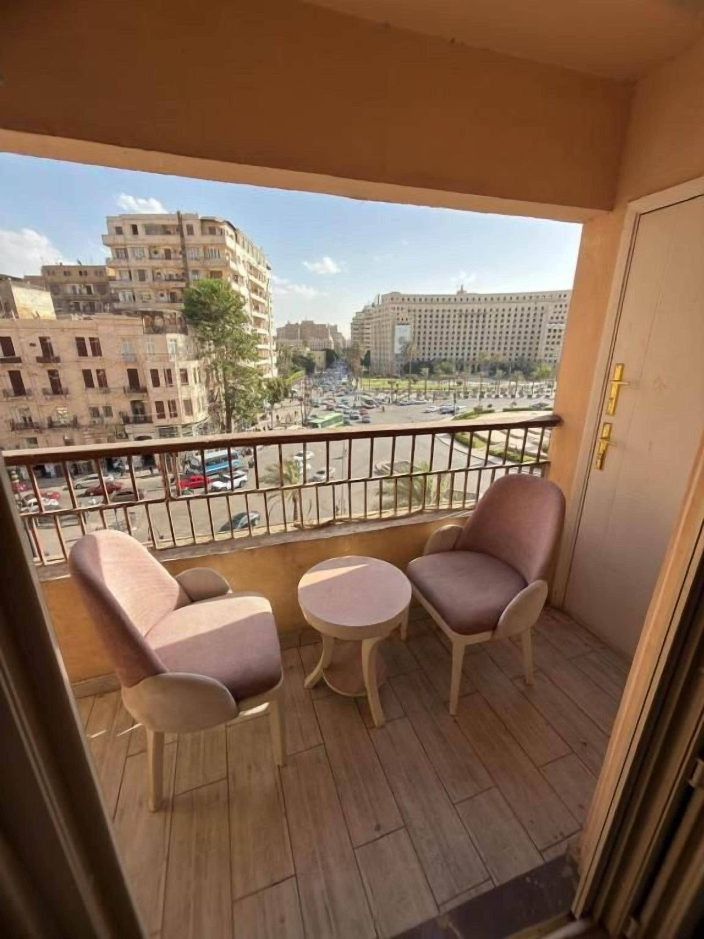 A balcony with two chairs and a small table, overlooking a busy street with buildings and parked cars.
