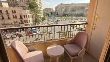 A balcony with two chairs and a small table, overlooking a busy street with buildings and parked cars.