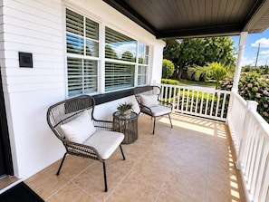 A covered porch with two wicker chairs and a small table, white railings, and a view of greenery.