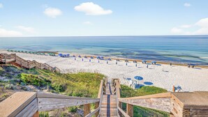 Une plage avec un escalier en bois descendant jusqu'au sable, des rangées de parasols bleus et une vue dégagée sur l'océan.