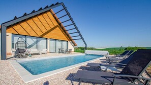 A modern building with a sloped roof and large glass windows, adjacent to a swimming pool with lounge chairs and a hanging chair.