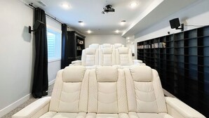 A home theater room with a row of white leather recliner seats, a ceiling-mounted projector, and a wall-mounted TV. - Cozy 6 bedroom home minutes from Rigby Lake (Rigby)