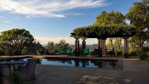 A pool area with green lounge chairs, a pergola, and a view of the mountains.