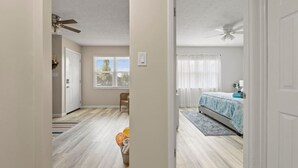 Un dormitorio con cama, ventilador de techo, ventana con persianas y puerta que da a otra habitación.