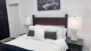Una cama bien hecha con ropa de cama blanca y azul marino, dos almohadas con estampado blanco y negro, un cabecero de madera oscura, un cuadro de un avión colgado en la pared y lámparas de noche a cada lado.