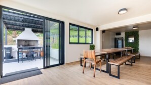 A modern outdoor dining area with wooden tables and chairs, a grill, and a view of a garden through large sliding glass doors.