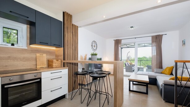 A modern kitchen with a built-in oven, a dining area with a wooden table and bar stools, and a balcony with outdoor seating.