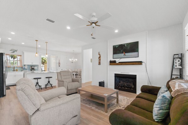 Una sala moderna con un sofá, un sillón y una mesa de centro. Hay una chimenea, un televisor montado en la pared y un ventilador de techo.