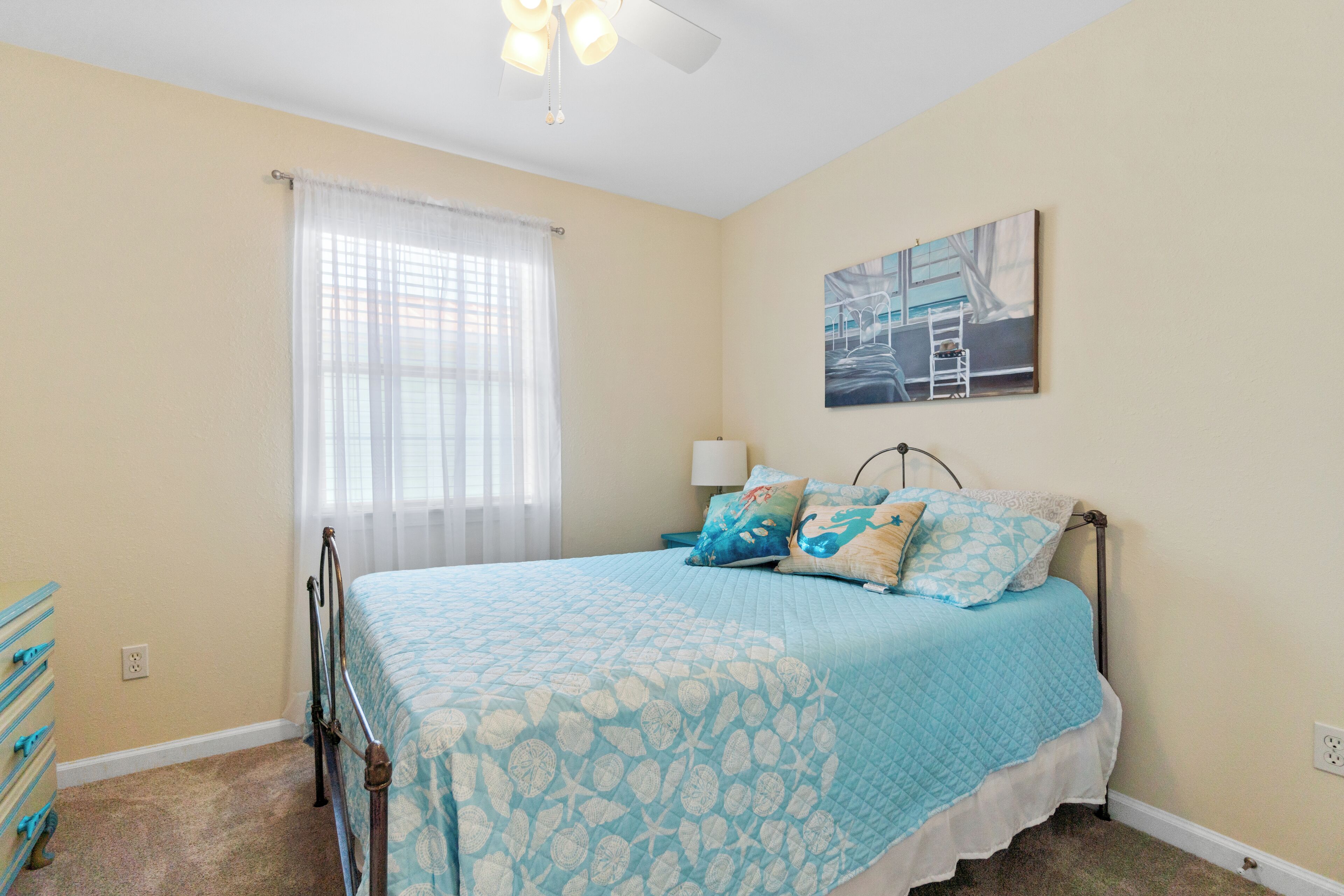 A bedroom with a bed, a nightstand, a lamp, a painting, and a window with curtains.