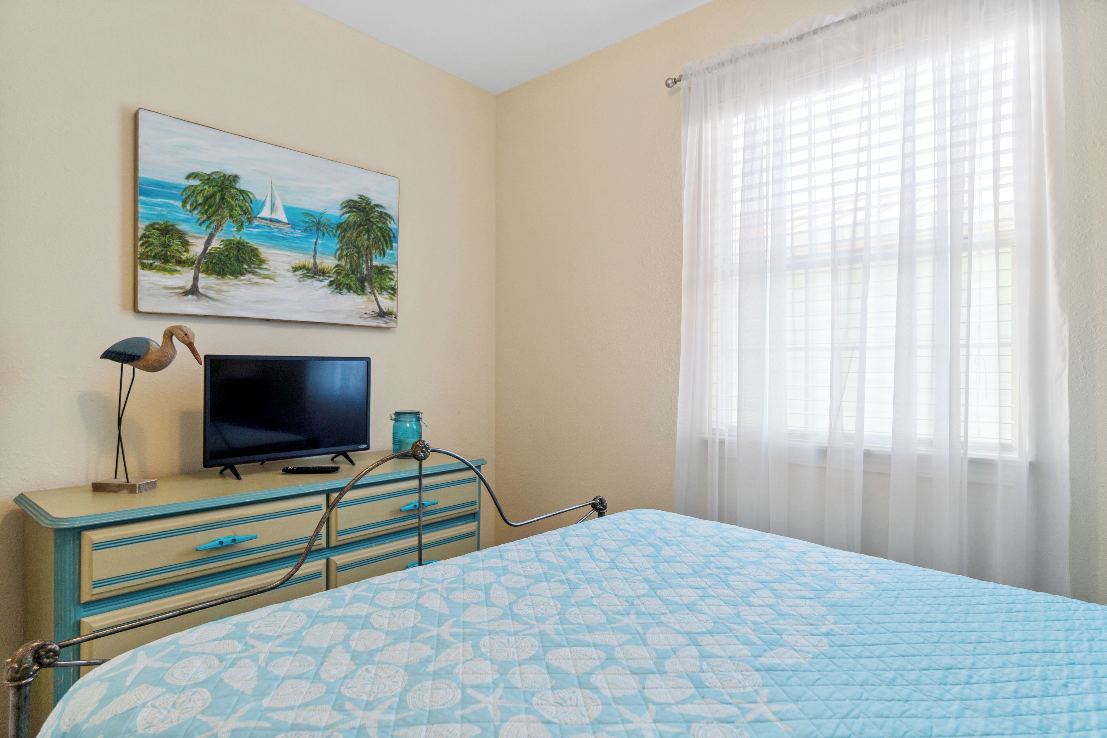 A bedroom with a bed, a television on a dresser, a painting of a beach scene, and a window with sheer curtains.