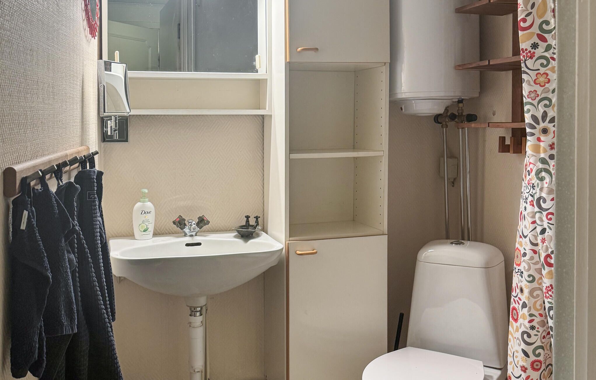 Un baño pequeño con un lavamanos blanco, un inodoro y una ducha. Hay un perchero con toallas oscuras y una botella de jabón Dove.