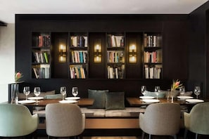 A modern dining area with a long table, cushioned seating, and bookshelves with books and wall-mounted lights. - Hotel Casa Volver (Barcelona)