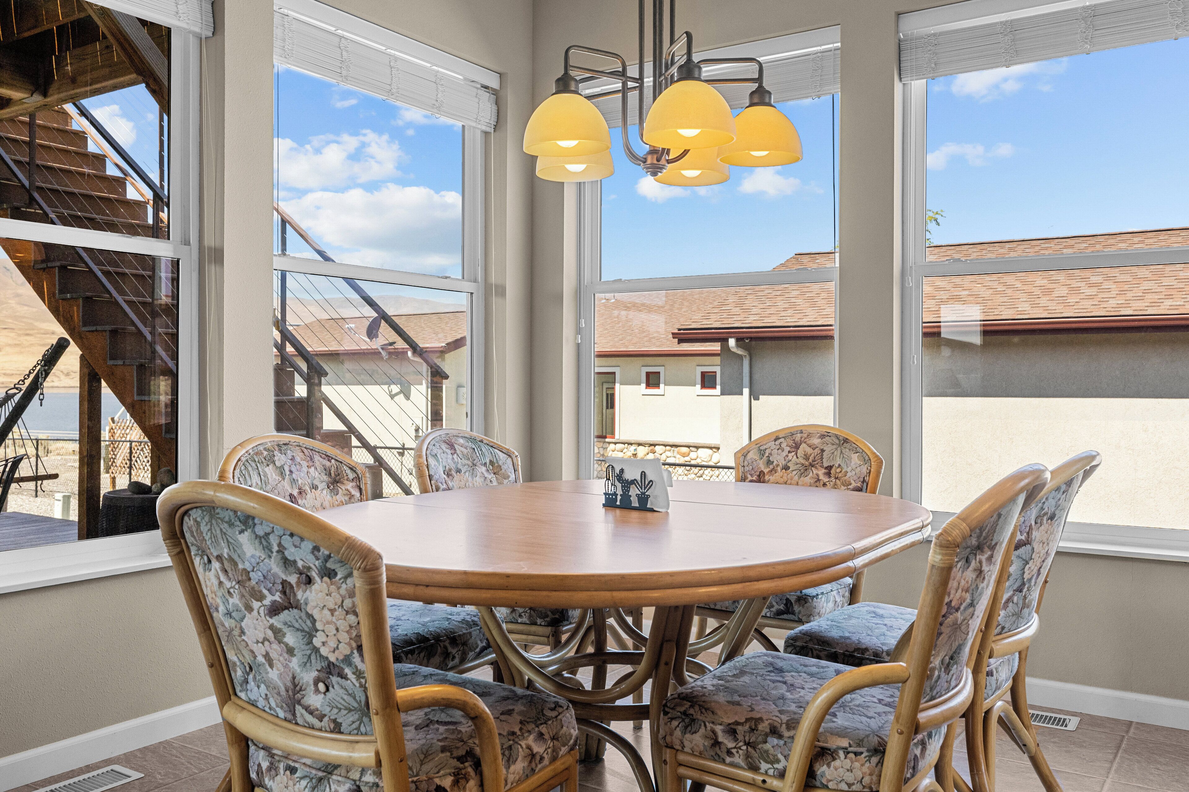 A dining area with a round table and floral-patterned chairs, large windows with a view of a building and a staircase, and a hanging light fixture.