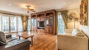 A spacious living room with a herringbone wood floor, a large wooden cabinet with glass doors, a ceiling fan, and a large wall clock.