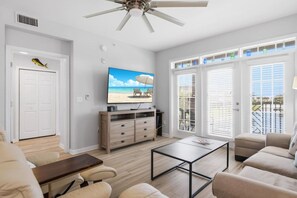 A living room with a television, a ceiling fan, a wooden cabinet, and a large window with blinds.