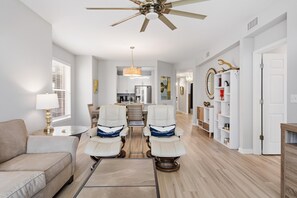 Amplia sala de estar con ventilador de techo, un sofá, dos sillones reclinables, una mesa de comedor y una cocina al fondo.