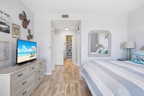 A bedroom with a television, a bed with striped bedding, a mirror, and a nightstand with a lamp.