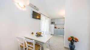Een compacte keuken met een magnetron, spoelbak en kastjes. Een eethoek met een tafel en stoelen is zichtbaar, en er staat een vaas met bloemen op de vloer.