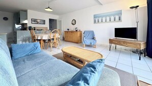 A modern living room with a sofa, a dining table with chairs, a TV stand, and a kitchen in the background.