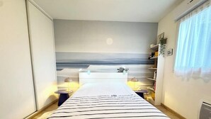 A bedroom with a bed, bedside tables, a nightstand, a window with curtains, and a wall-mounted shelf with books and a plant.