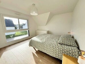 Une chambre avec une grande fenêtre, un lit recouvert d’une literie à motifs, une table de chevet et un luminaire suspendu.