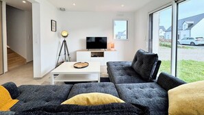 Un salon moderne avec un canapé d’angle, un téléviseur à écran plat et une grande fenêtre donnant sur l’extérieur.