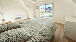 Une chambre avec un grand lit, un luminaire au plafond, une fenêtre donnant sur des maisons et une armoire blanche.