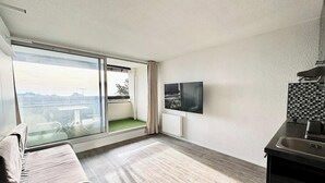 A modern apartment with a large sliding glass door leading to a balcony, a flat-screen TV mounted on the wall, and a kitchenette area. - Studio Confort - Vue sur Mer - Balcon (Guidel)