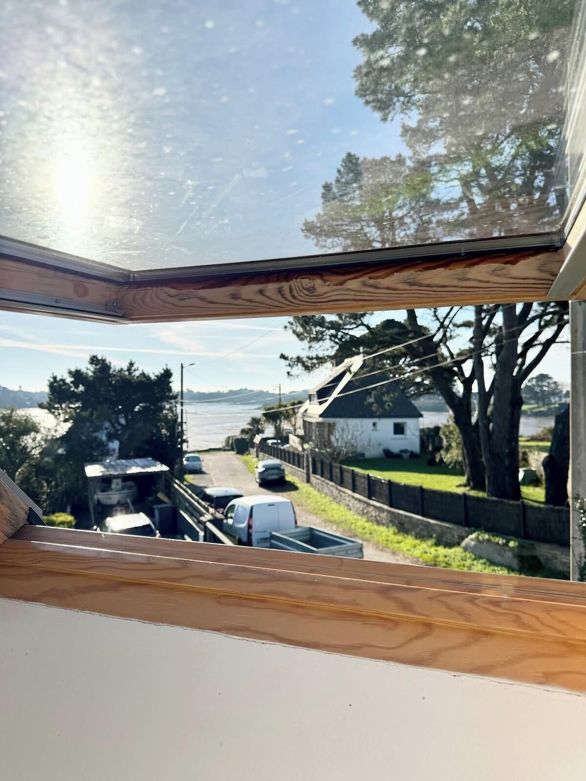 Une vue depuis une fenêtre à cadre en bois, donnant sur une rue bordée de voitures garées et de maisons, et sur une étendue d’eau au loin.