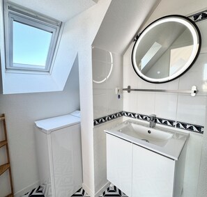Een kleine, witte badkamer met een raam, een ronde spiegel, een wastafel en een handdoekhouder.