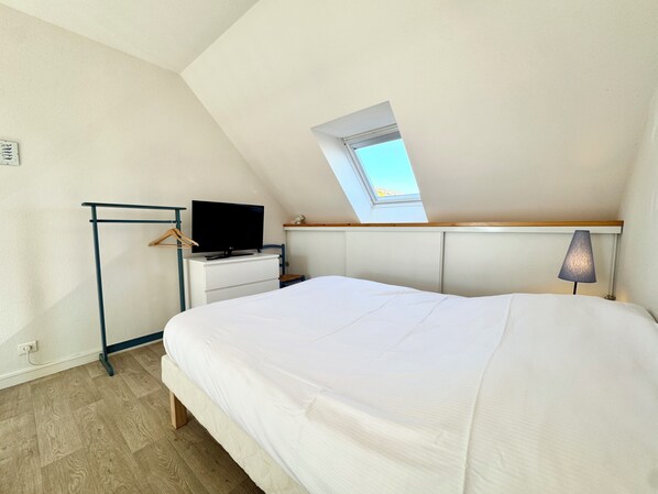 A bedroom with a sloped ceiling, a skylight, a bed, a TV on a dresser, and a lamp.