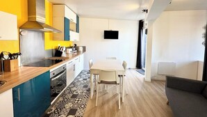 A modern kitchen with a dining area, a flat-screen TV mounted on the wall, and a sofa in the living space.